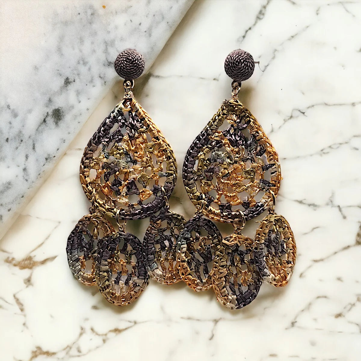 A pair of large, metallic, shimmering teardrop chandelier earrings with intricate gold and black woven patterns displayed on a marble surface.