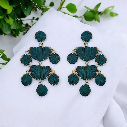 A pair of green and gold chandelier earrings featuring circular and semicircular elements, exuding bohemian elegance, displayed on white fabric with green leaves in the background.
