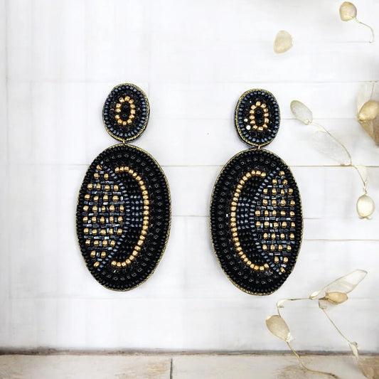 A pair of black and gold mixed abstract oval drop earrings with glass beadwork, displayed against a light-coloured background with dried decorative branches on the right.