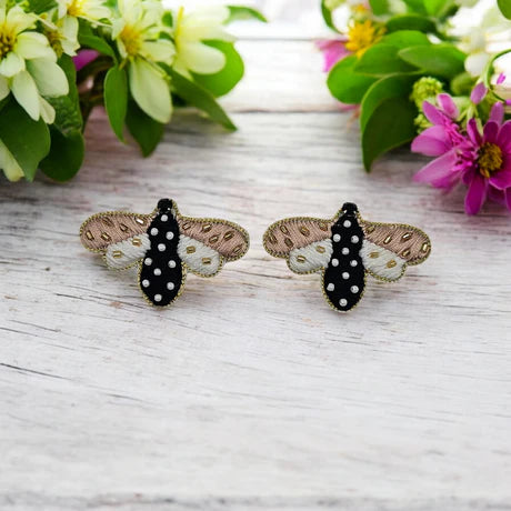 Two embroidered bee earrings with black, white, and gold details rest on a white wooden surface, surrounded by green leaves and colourful flowers. These unique stud earrings make a charming accessory.