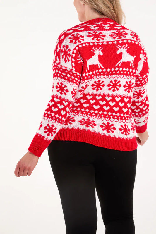 Person wearing a red and white festive jumper featuring reindeer, snowflake, and heart patterns, paired with black trousers, standing against a plain background.