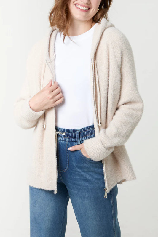 A person wearing a white T-shirt, blue jeans, and the Super Soft Stone Hoodie—a sustainable hoodie—stands with one hand in their trouser pocket and the other holding the hoodie.