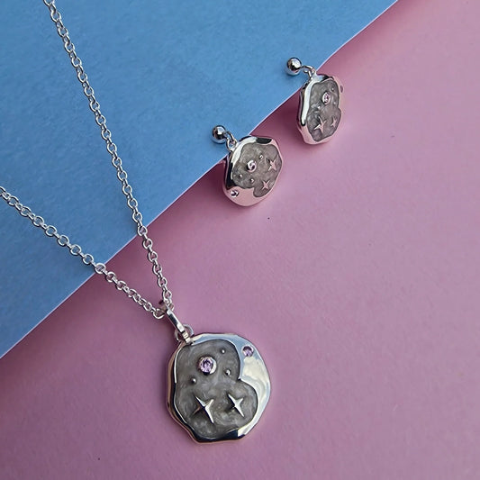 Silver necklace and celestial earrings set featuring textured circular pendants, delicate star and swirl engravings, and pink gemstones, presented on a blue and pink background.