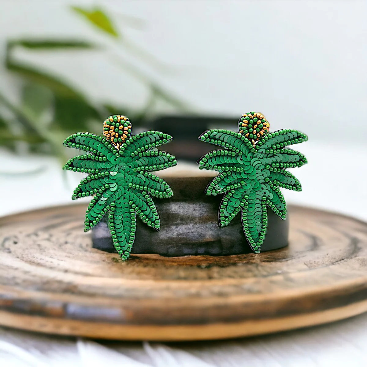 A pair of green beaded earrings with a tropical leaf design and small orange bead accents, displayed upright on a round wooden surface.