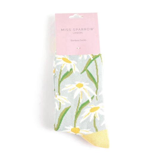 A pair of vegan socks, the Duck Egg Oxeye Daisies Socks feature a white daisy and green leaf pattern, yellow toe, and pink packaging labelled Miss Sparrow London.
