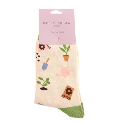 A pair of cream-coloured, British-designed Miss Sparrow bamboo socks with a gardening-themed pattern, featuring plants, watering cans and tools. The vegan-friendly socks have green toes.