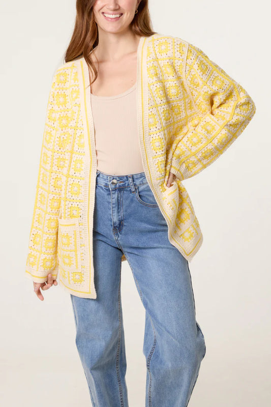 Woman wearing a yellow and white patchwork cardigan over a beige top and blue high-waisted jeans, standing against a plain light background.