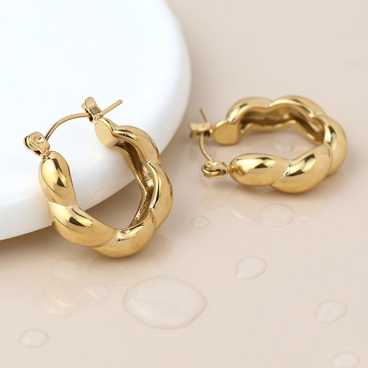 A pair of waterproof gold-plated hoop earrings with a twisted design shown on a beige surface with water droplets, beside a white circular object.