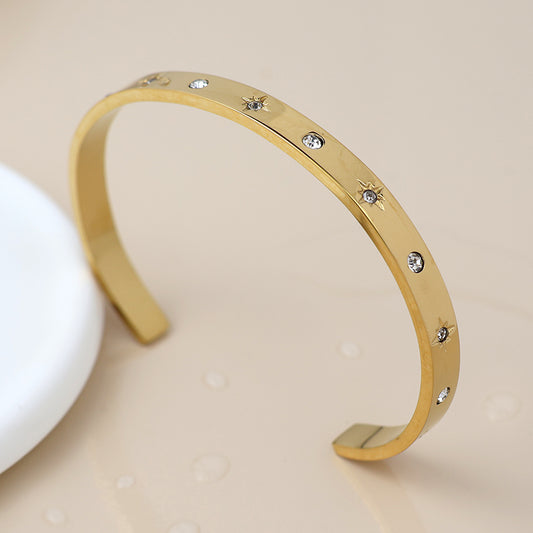 Gold crystal-studded bangle featuring clear gemstones evenly spaced across its surface, shown on a beige background with water droplets.