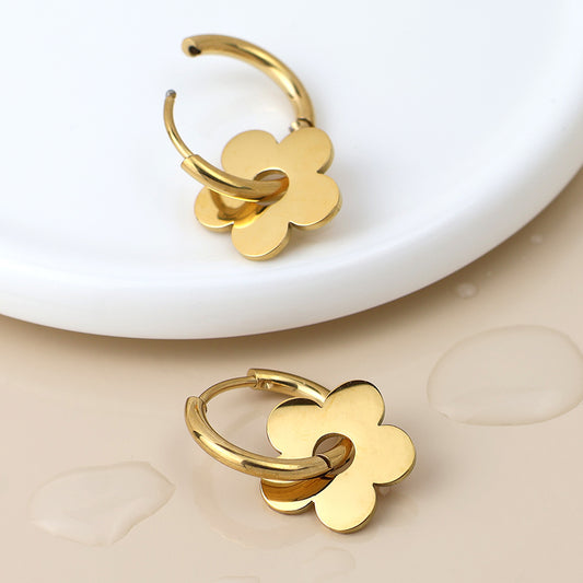 Two gold-plated hoop earrings featuring flat, flower-shaped charms are shown on a beige surface beside a white ceramic dish and water droplets—stylish, waterproof earrings ideal for everyday sophistication.