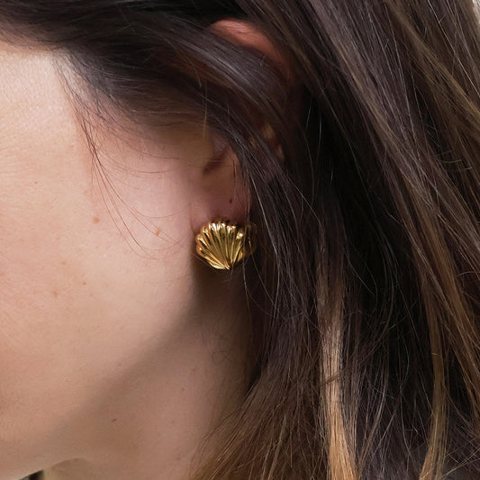 Close-up of a person's ear adorned with a gold clam shell earring, ideal as seaside jewellery; brown hair partially visible.