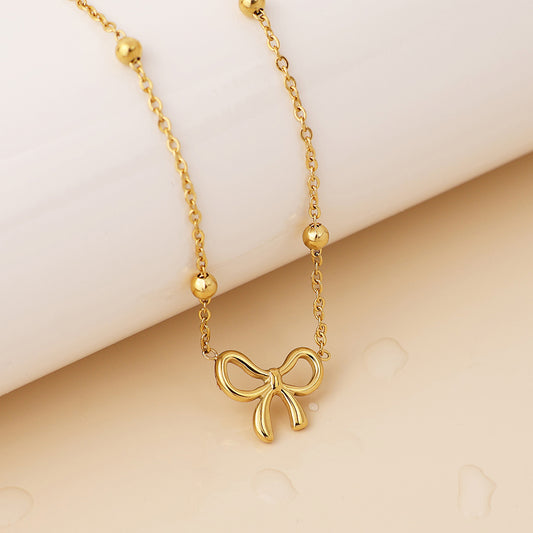 Gold-plated necklace with a delicate bow pendant and small round beads, shown on a light, glossy surface.