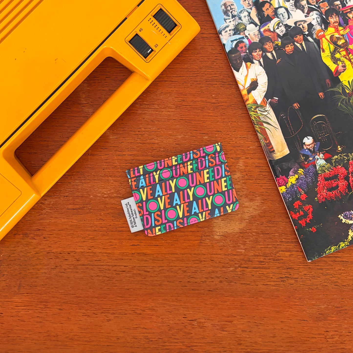 A small, colourful retro cardholder featuring the phrase All You Need Is Love rests on a wooden surface beside a yellow portable device and a partially visible album cover.