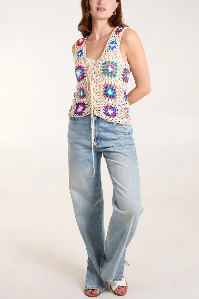 A fashion-forward woman wears a vibrant patchwork crochet waistcoat over a white top, paired effortlessly with light blue jeans and sandals.
