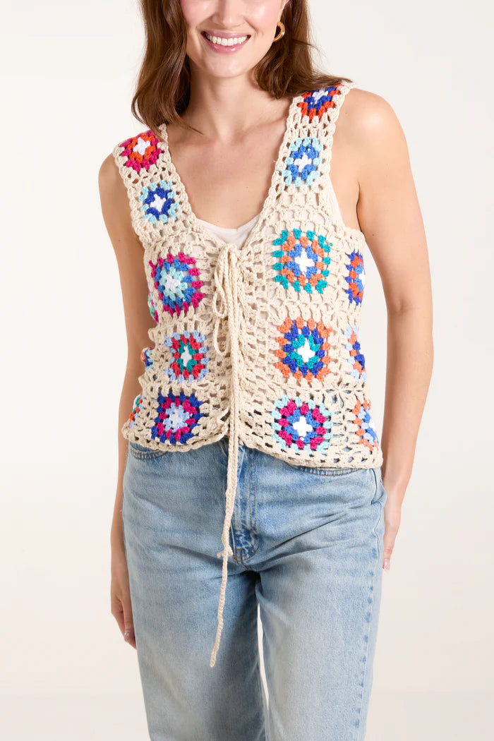 A fashion-forward woman wears a vest top, featuring colourful patchwork crochet with geometric patterns, over a white shirt and light blue jeans.
