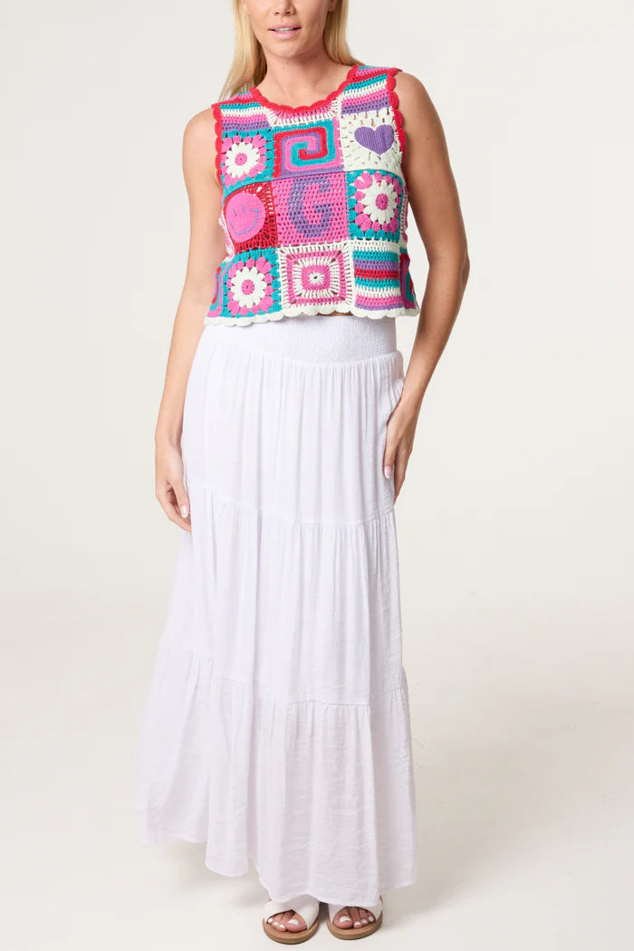 A woman in a colourful, 70s-inspired crocheted sleeveless top and a long, white tiered skirt stands against a simple light background.