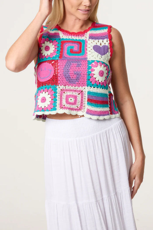 A person sporting a colourful retro patchwork crochet vest adorned with assorted patterns and symbols, teamed with a white skirt, stands before a plain background, radiating vintage charm.