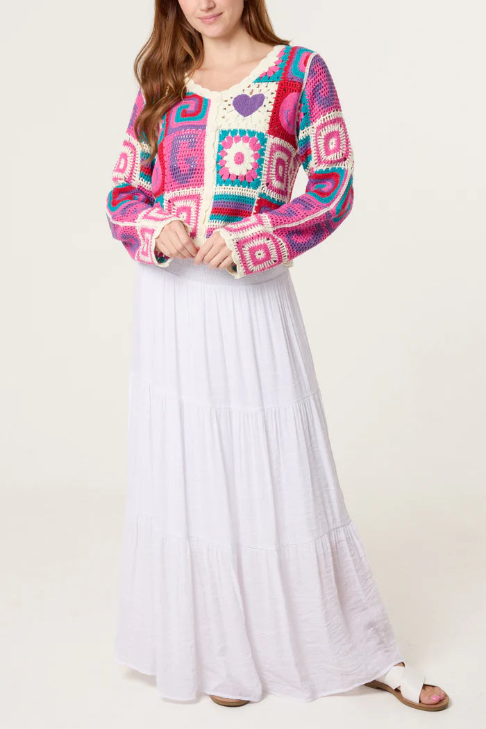 A person wearing a colourful retro patchwork crochet cardigan featuring square and heart patterns, paired with a long, white tiered skirt and white sandals.