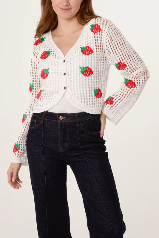 A person dressed in a white, vintage cardigan adorned with red strawberry embroidery, dark jeans, and a white top poses with one hand on their hip.