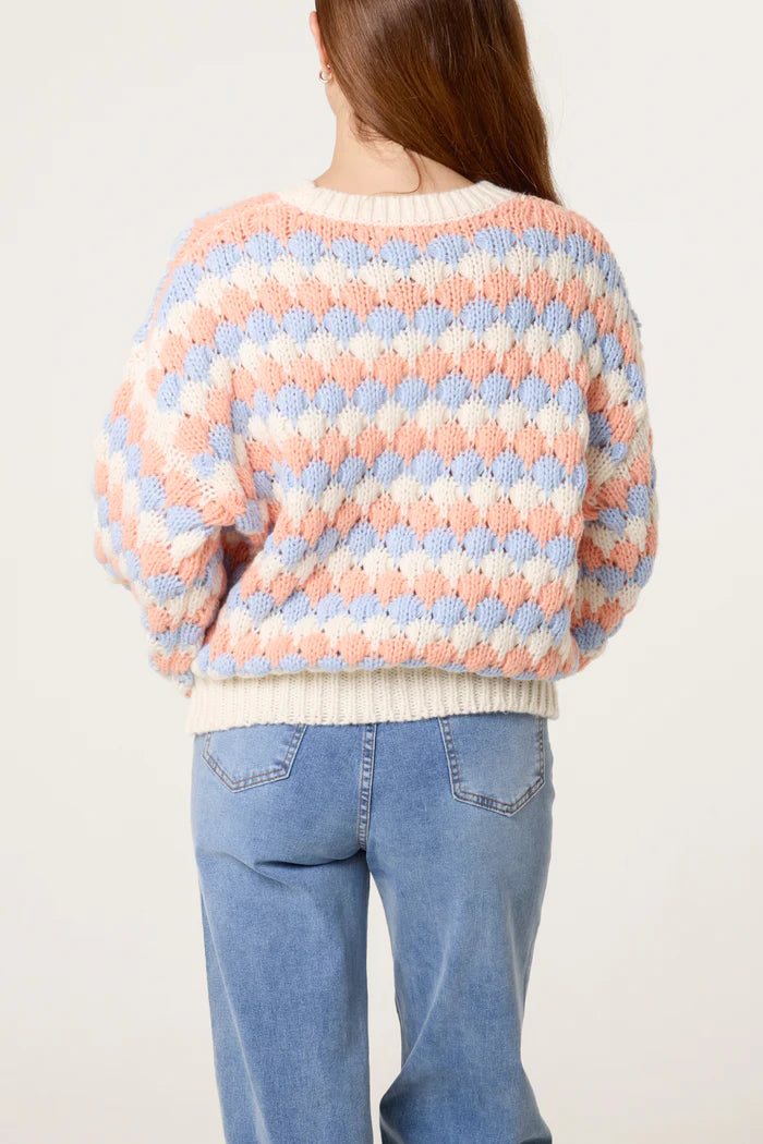 A person with long brown hair is seen from behind, wearing a vegan-friendly jumper in blue and peach chequered knit and light blue jeans, set against a plain background.