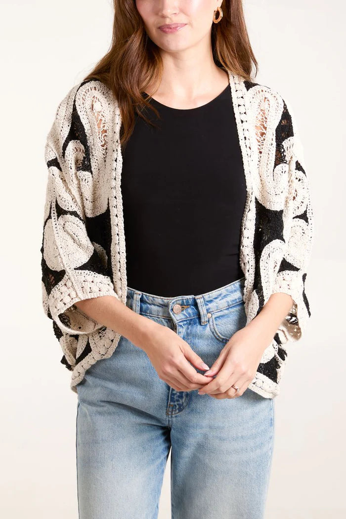 The person is wearing a black top, light blue jeans, and an open beige and black patterned cardigan in a batwing style, reminiscent of a paisley crochet kimono.