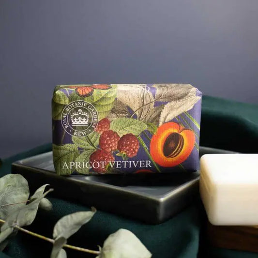 A botanical-patterned Apricot Vetiver Soap bar rests on a dark tray beside a white vegan soap and vibrant green foliage.