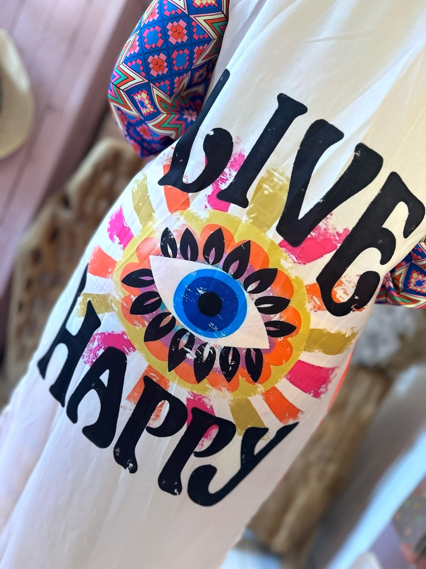 A colourful, vegan-friendly garment for your summer wardrobe, featuring bold “LIVE HAPPY” text and a large blue eye surrounded by vibrant patterns.