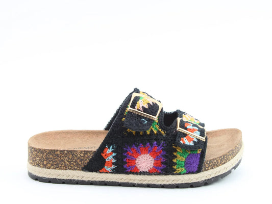 A black slip-on sandal with a cork sole, open toe, two buckled straps, and a colourful stitched upper featuring crochet flower patterns. Includes a contoured footbed for added comfort.