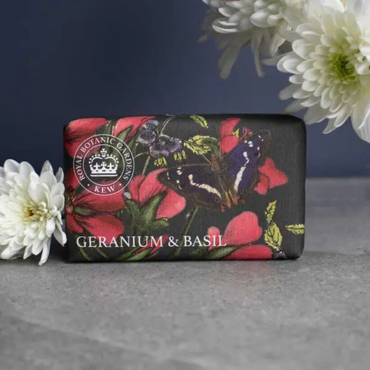 A Kew Gardens Geranium and Basil Soap bar, adorned with floral and butterfly illustrations, sits on a grey surface beside white flowers; this vegan-friendly soap brings elegance to your daily routine.