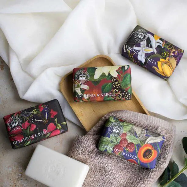 Four wrapped bars of vegan-friendly soap with botanical designs are arranged on a wooden tray and towel, beside an unwrapped white bar of soap on a neutral surface.