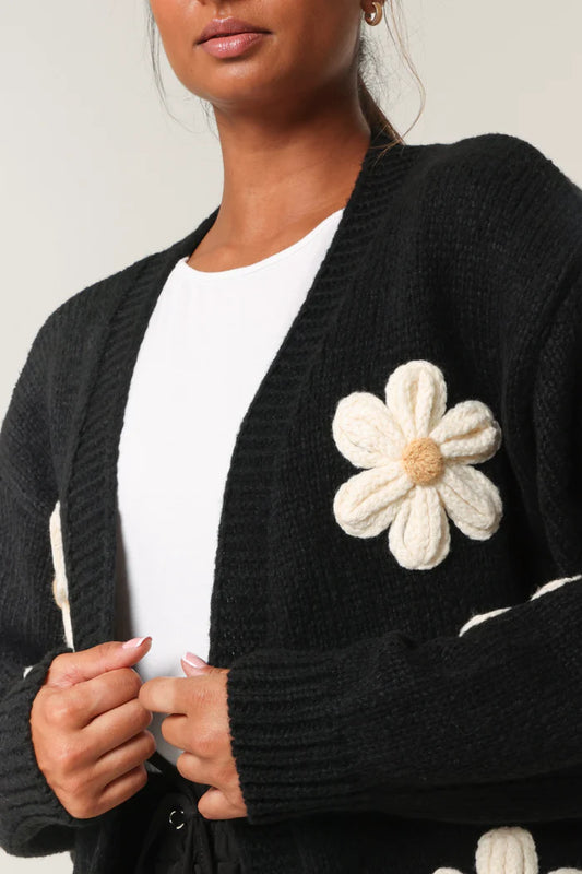 A woman wears a hand-knitted cardigan adorned with large cream-coloured flower appliqués over a white top, showcasing sustainable style.