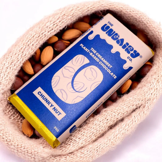A blue and white Chunky Nut no-dairy chocolate bar with a vegan twist is surrounded by assorted nuts and resting on beige knitted fabric.