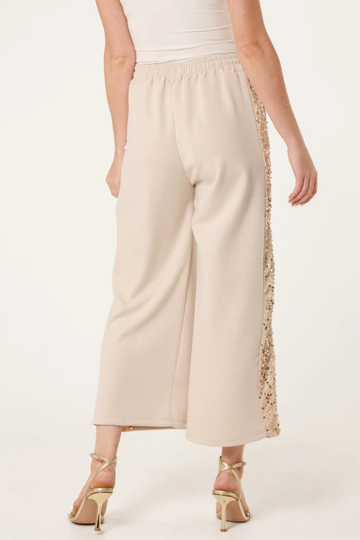 A person wearing wide-leg, cream-coloured sequin velvet joggers from the Italian collection, paired with a white top and strappy heeled sandals, seen from the back.