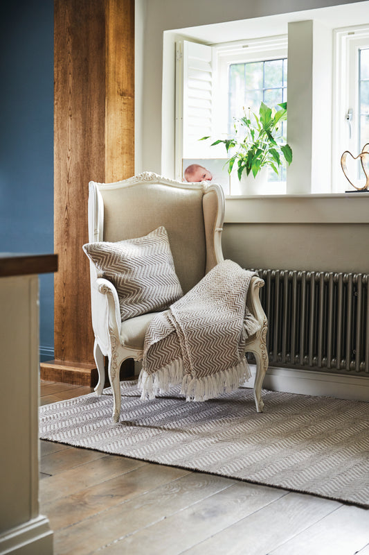 A decorative armchair with eco-friendly cushions made from recycled plastic bottles and a throw sits by a window. Light streams in, illuminating the wooden floor and the potted plant on the sill.