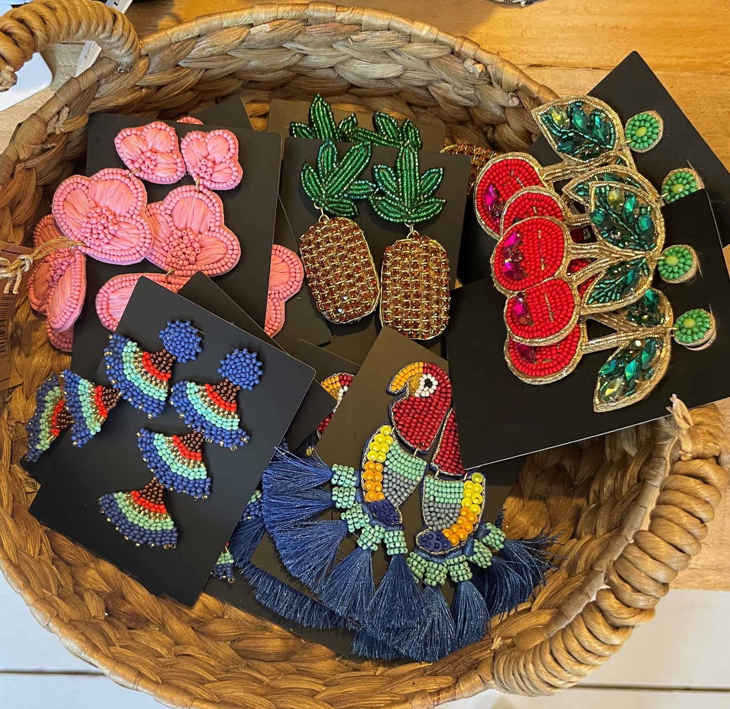 A woven basket filled with Carnation Pink Earrings and other colourful beaded designs—flowers, rainbows, pineapples, cherries and a parrot—all statement jewellery pieces displayed on black cards.