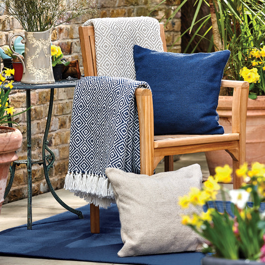Wooden outdoor chair with blue and patterned cushions, draped with a sustainable throw made from recycled plastic bottles, beside a metal table and surrounded by potted plants and yellow flowers.