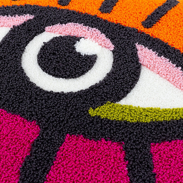 Close-up of a colourful textured rug showcasing a stylised eye design with black, white, pink, orange, and green sections, complemented by a vibrant colourful throw cushion for added flair.