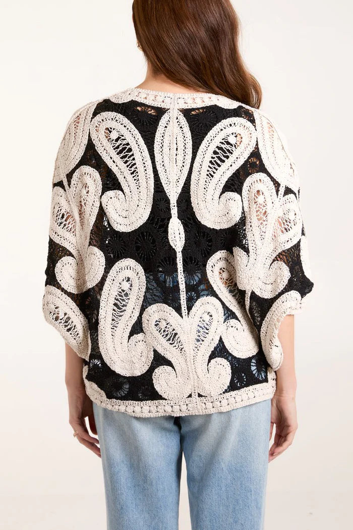 An individual dressed in a black and white crocheted jumper featuring an intricate batwing style pattern and light blue jeans, viewed from behind.