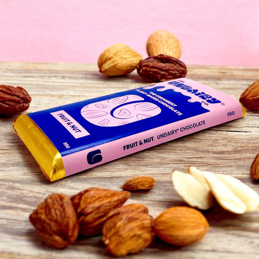 A Fruit and Nut Chocolate bar in pink and blue packaging is surrounded by whole and shelled nuts on a wooden surface with a pink background, showcasing this vegan chocolate treat.