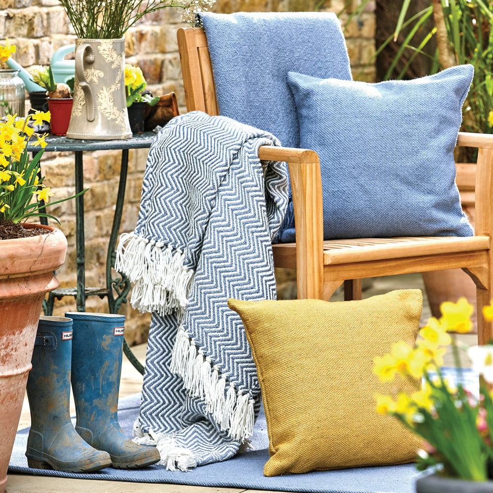 Wooden chair with blue cushions and an eco-friendly herringbone throw on a terrace, next to potted daffodils and muddy blue wellies, with a watering can on a small table.