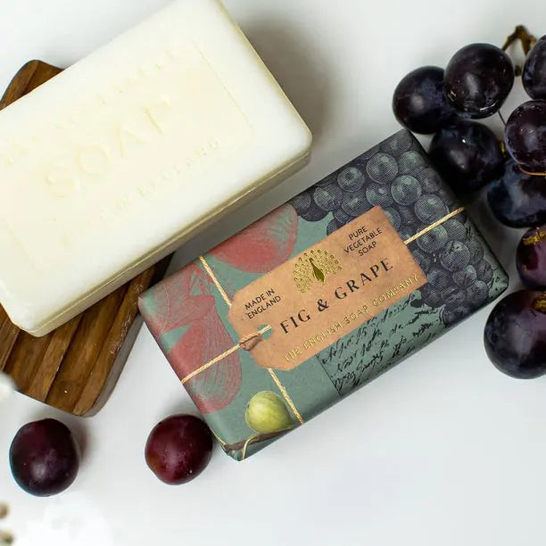 Two bars of vegan-friendly Anniversary Fig and Grape Soap, one unwrapped and one in decorative packaging, shown next to fresh grapes and a wooden soap dish.