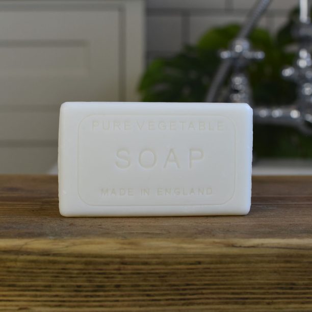 A white rectangular bar of vegan-friendly soap, labelled "Pure Vegetable Soap Made in England," sits on a wooden surface with a kitchen sink and green plant in the background.