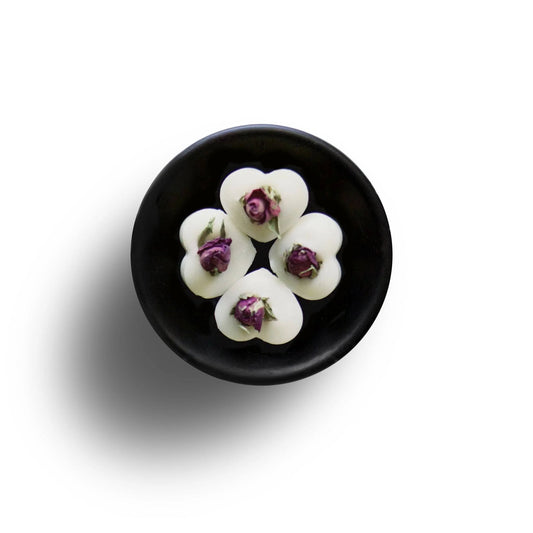 A black dish displays five white heart-shaped items, each adorned with a small pink rosebud, creating an elegant presentation of English Rose Soya Wax Melts. Perfect for infusing your space with a luxurious home fragrance.