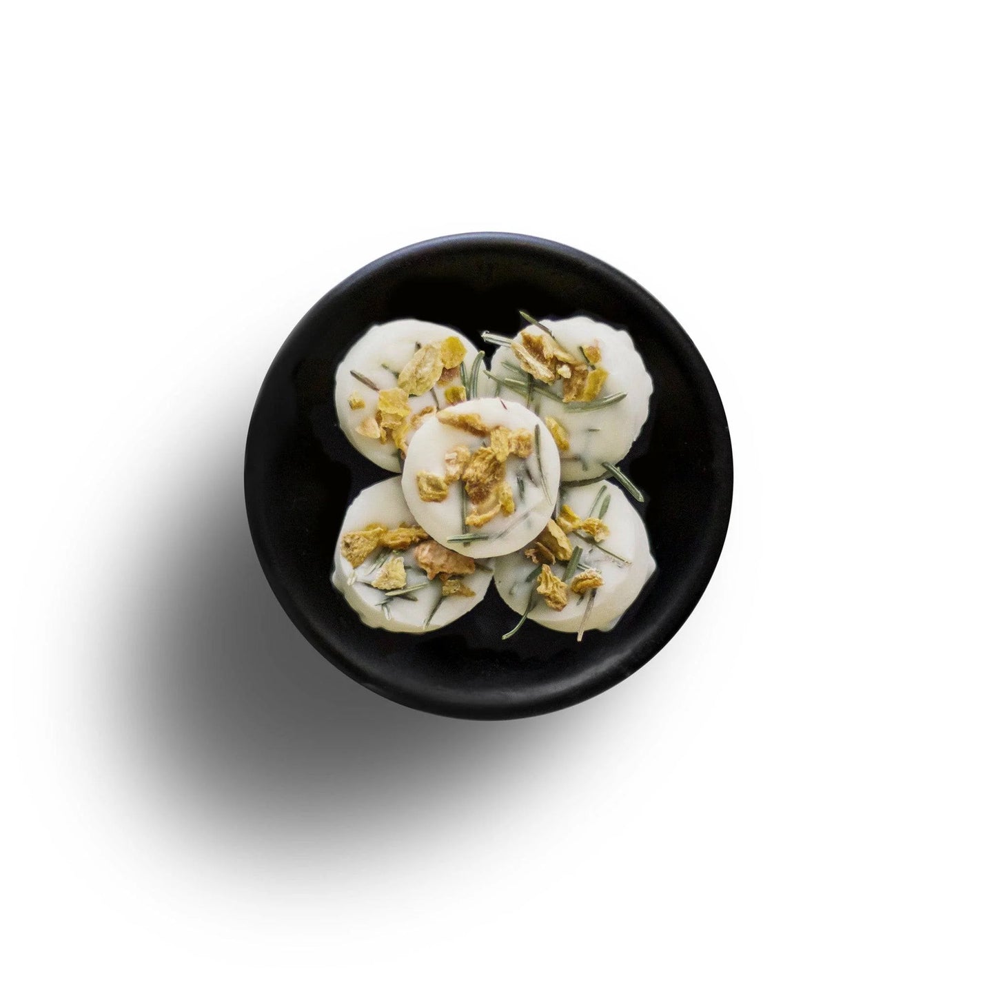 On a black plate, five white circular snacks are topped with crushed nuts and herbs, resembling eco-friendly wax melts. They rest elegantly against a pristine white backdrop.