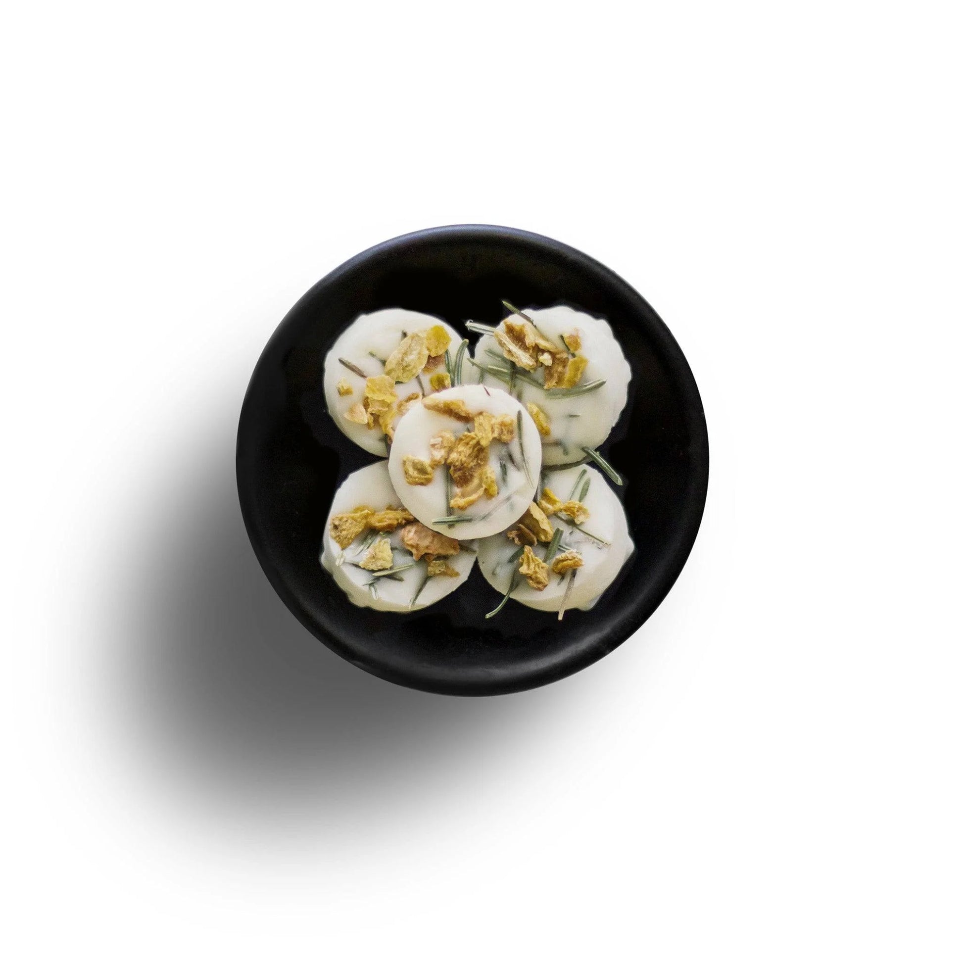 On a black plate, five white circular snacks are topped with crushed nuts and herbs, resembling eco-friendly wax melts. They rest elegantly against a pristine white backdrop.