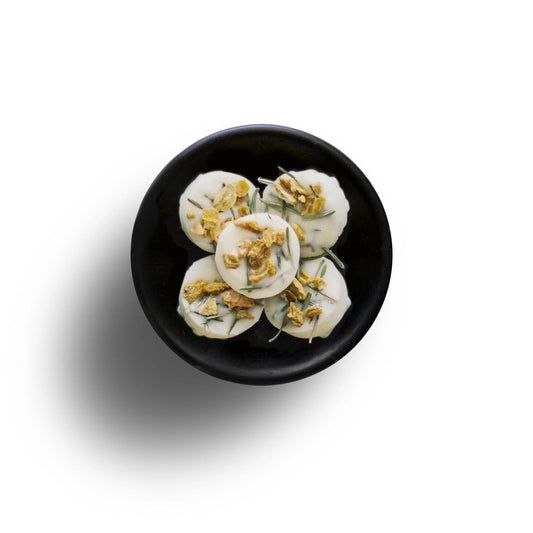 On a black plate, five white circular snacks are topped with crushed nuts and herbs, resembling eco-friendly wax melts. They rest elegantly against a pristine white backdrop.