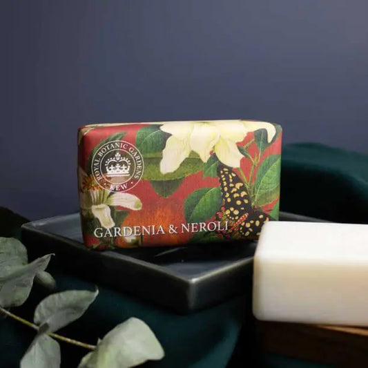 A bar of Gardenia and Neroli Soap in plastic-free packaging rests on a dark dish next to an unwrapped vegan soap bar and several eucalyptus leaves.