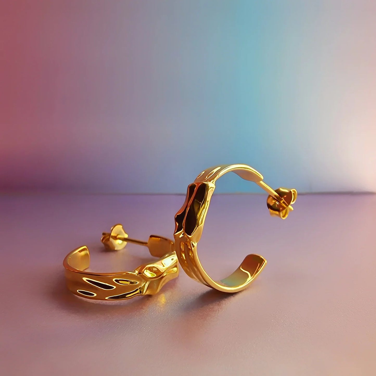 A pair of gold crescent hoop earrings with abstract, wavy designs shown on a smooth surface against a pastel gradient background.