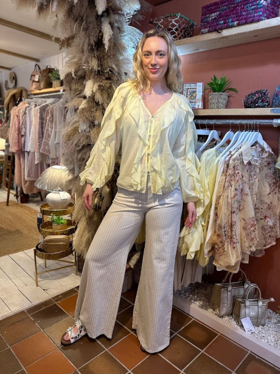 A woman stands indoors in a boutique, radiating feminine elegance in a light yellow ruffle-front blouse and wide-leg striped trousers, surrounded by clothing rails and stylish decorative items.
