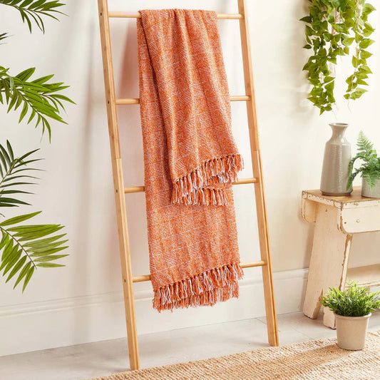 An orange and white Woven Cross Hatch Rust Throw with fringed edges hangs on a wooden ladder against a white wall, surrounded by green plants and neutral décor.